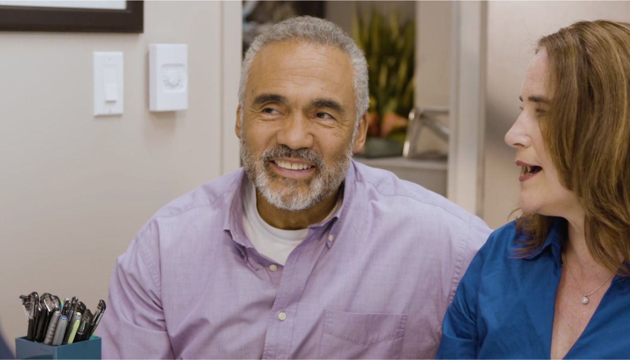 Older man smiling while sitting next to his partner.