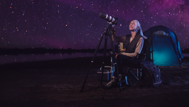 Woman wearing a black sleeveless dress sitting down on a chair underneath a starry sky. There’s a tent in the background and she is looking through a telescope at a beautiful starry purple night sky with a look of wonder on her face.